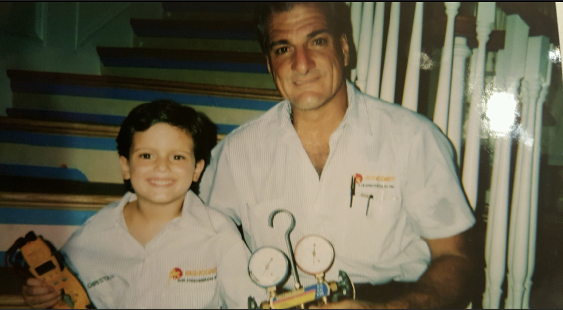 A smiling man and boy pose with an HVAC device on a staircase. Both wear work shirts.