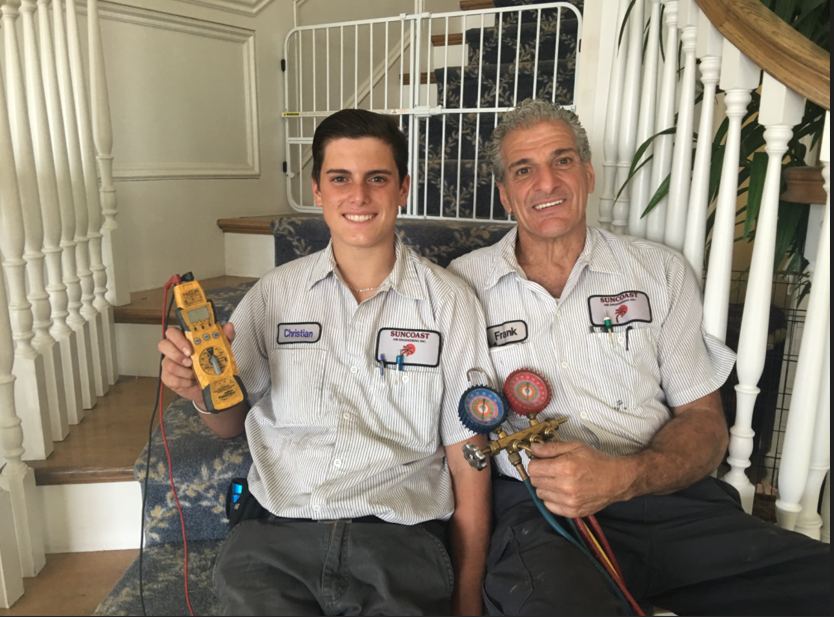 Two HVAC technicians sit on steps, smiling, holding tools. White gate, staircase, floral carpet.