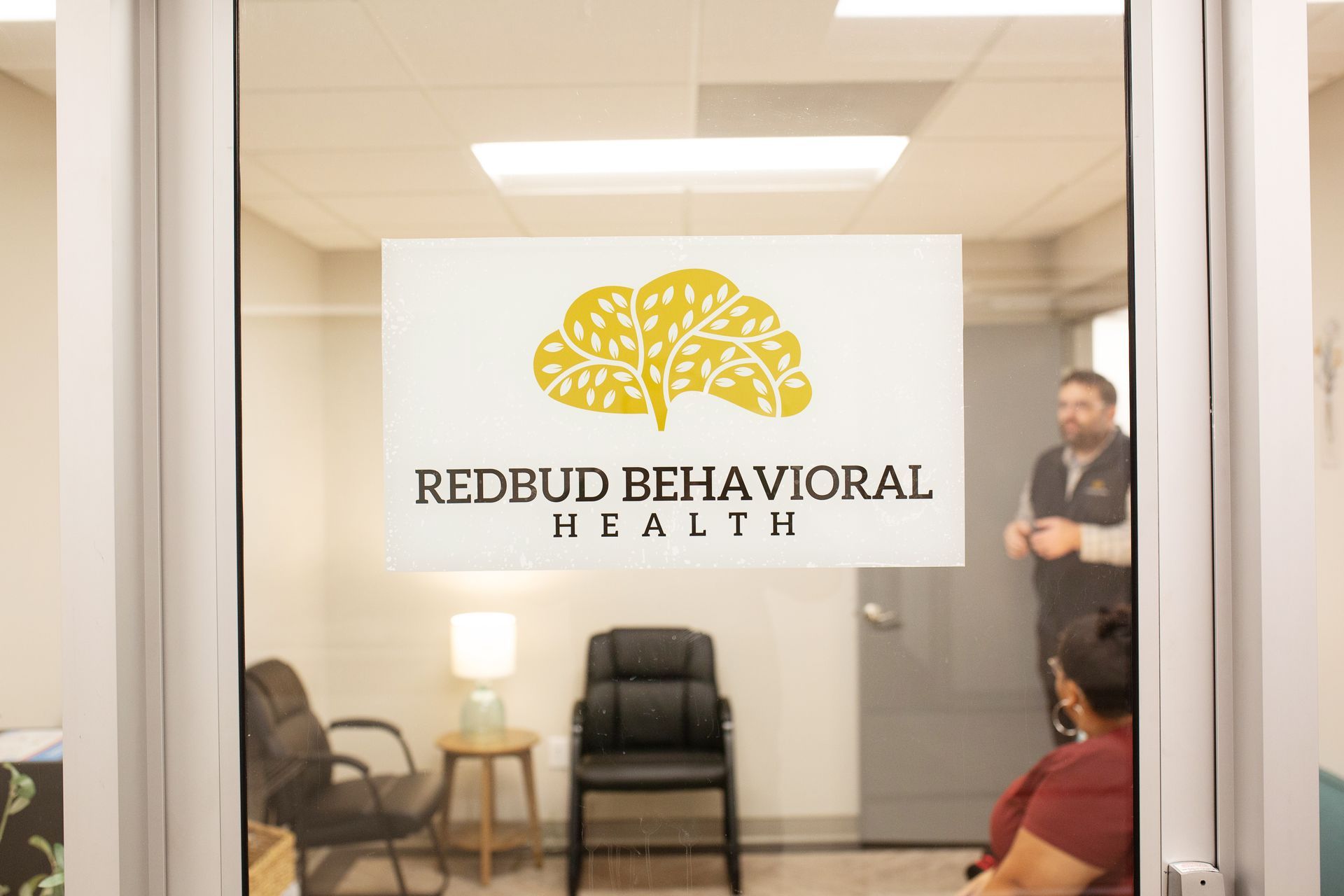 Through glass door: Redbud Behavioral Health logo above a waiting area with a person speaking.