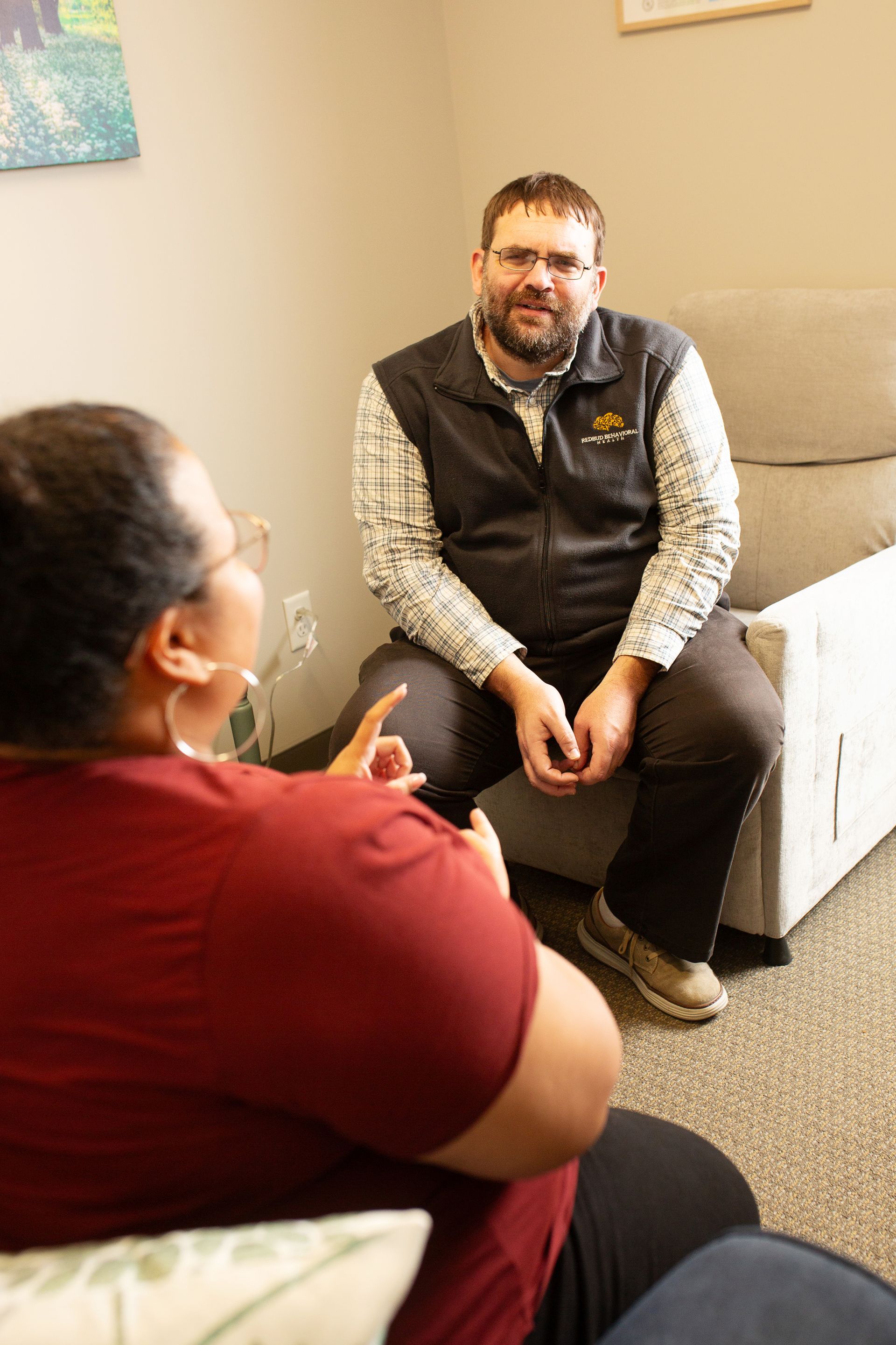 A person in glasses and a vest sits and listens to a person with their back to the camera.