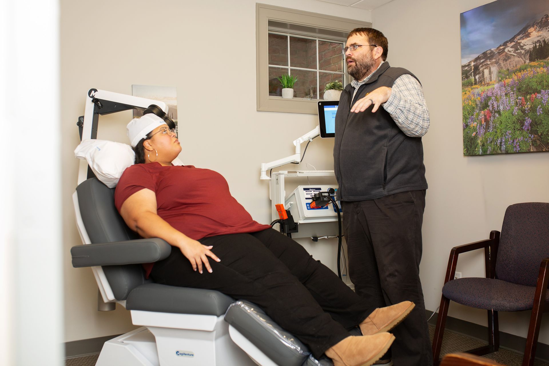 A medical professional explains something to a patient seated in a chair, with medical equipment in a clinic.