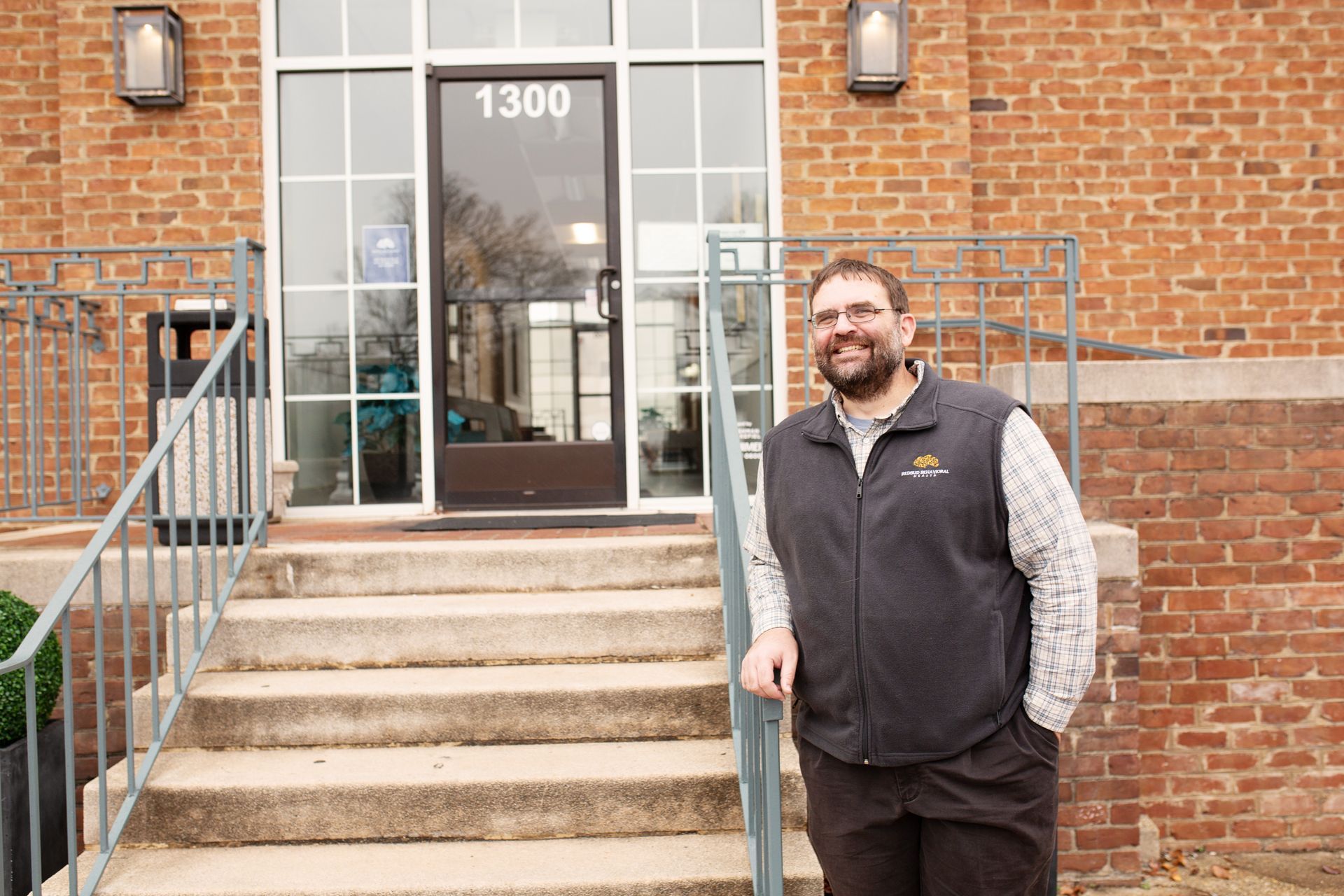 Man in vest smiles at camera outside building entrance with the number 1300.