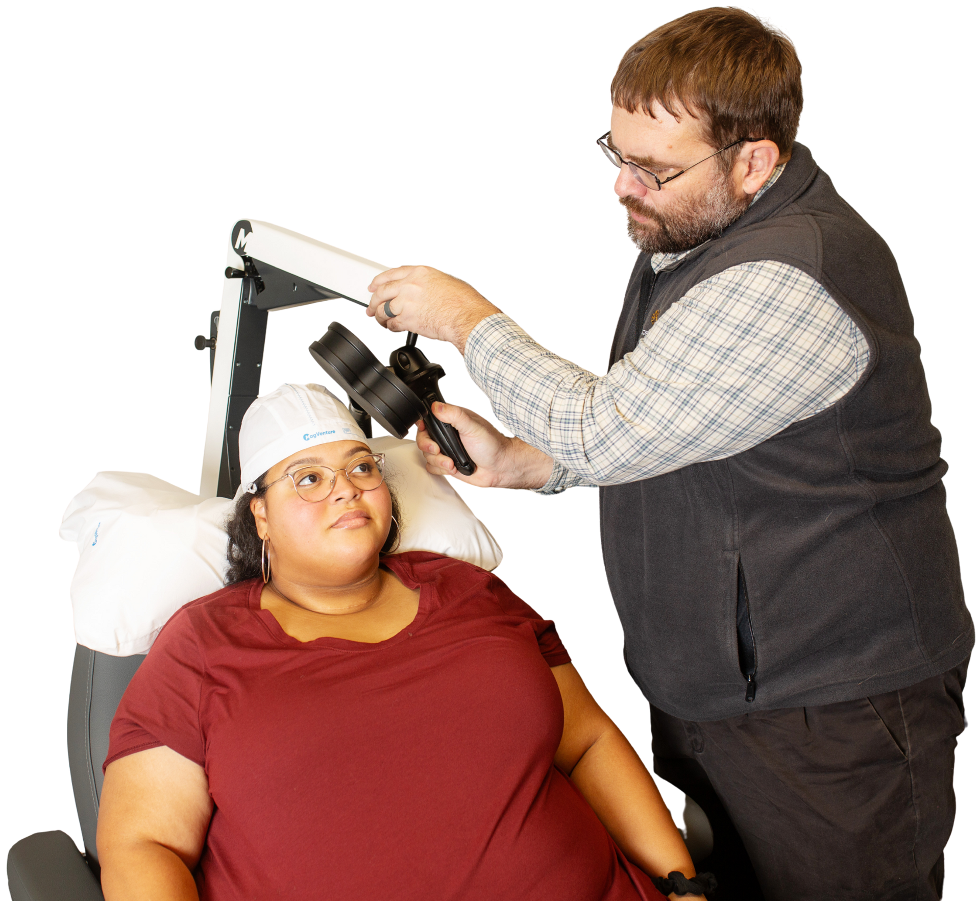 Person adjusts equipment on the head of another person seated in a chair, likely for a medical scan.