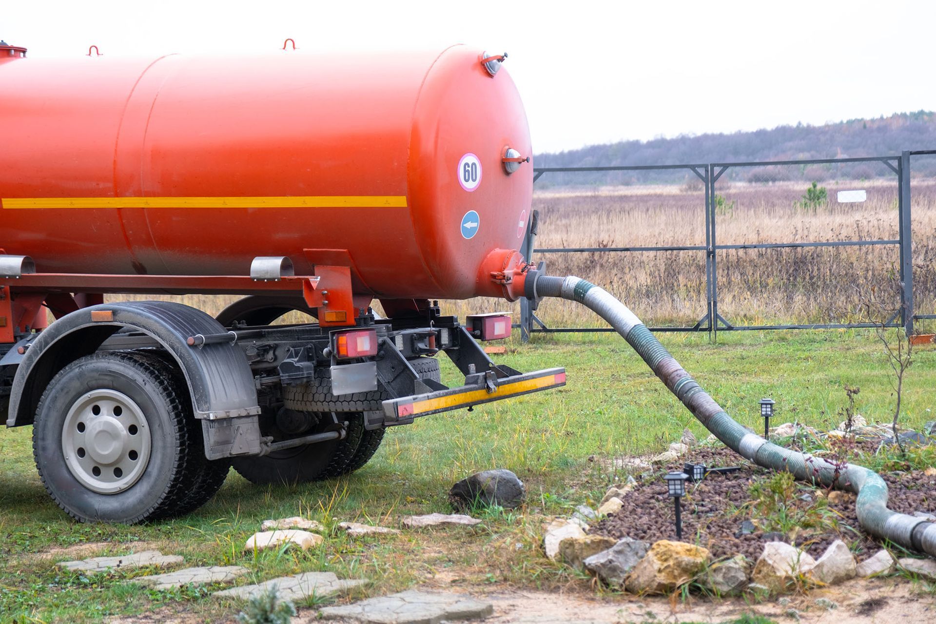 An orange tanker truck is parked on grass, connected by a hose to an intake pipe in the ground.