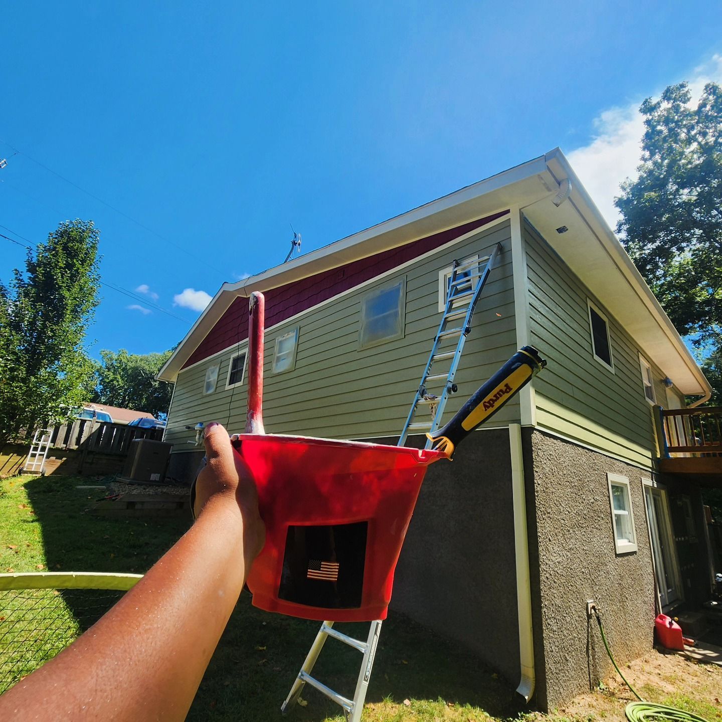 Person holding a red paint bucket in front of a house being painted; ladder propped against the house.