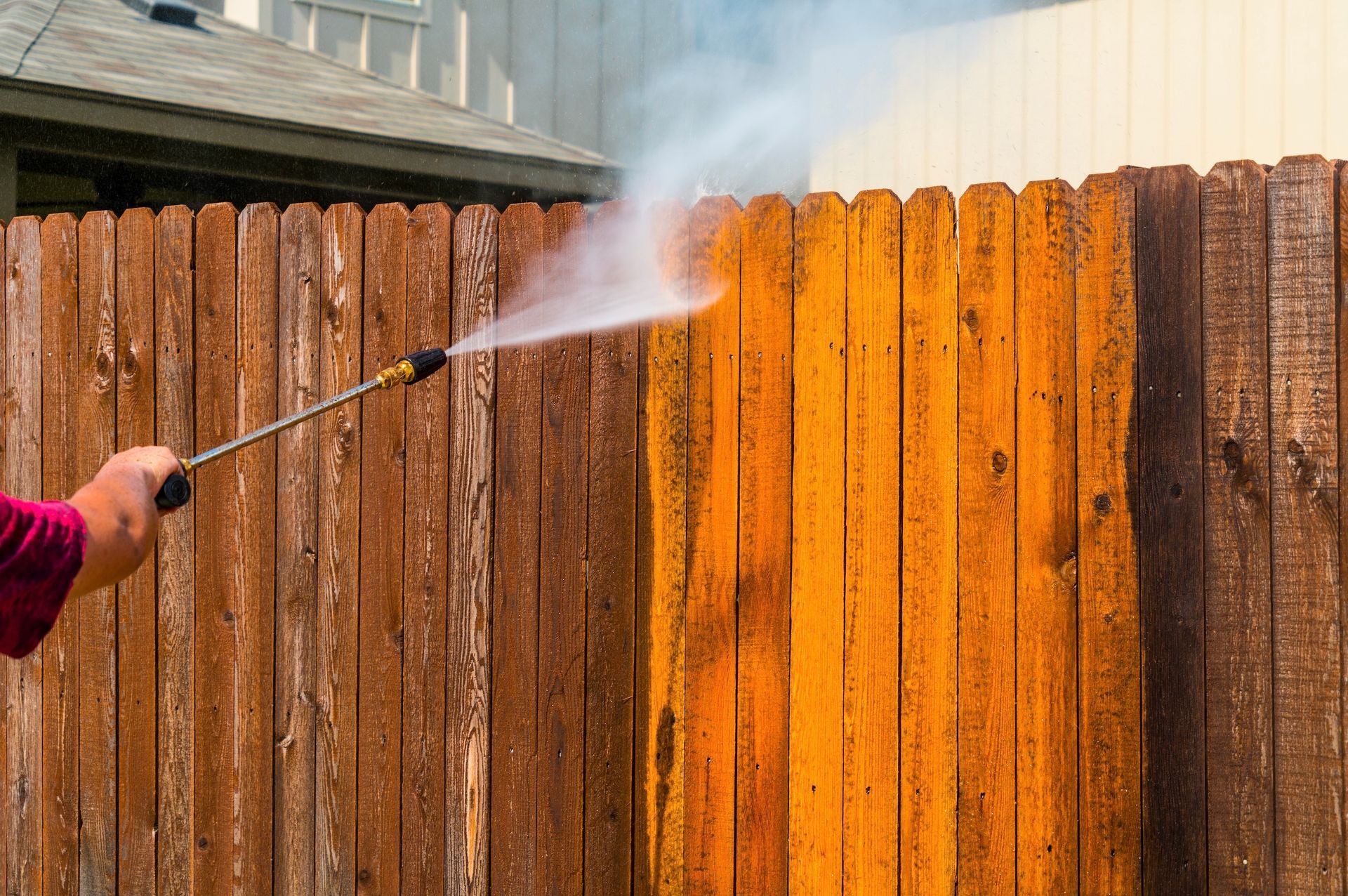 Person pressure washing a wooden fence, cleaning off dirt and grime.