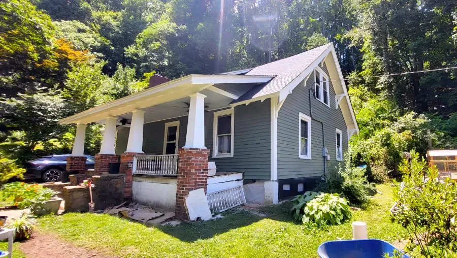Green house with white trim, porch, and a brick base, set in a wooded area.