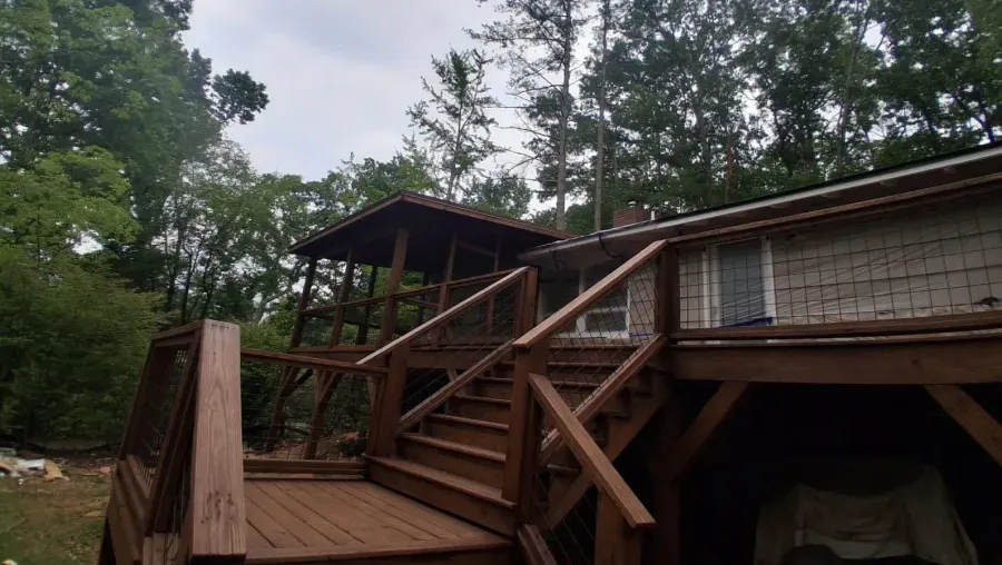 Wooden deck with stairs leading to a screened porch attached to a house. Overcast sky and trees in the background.