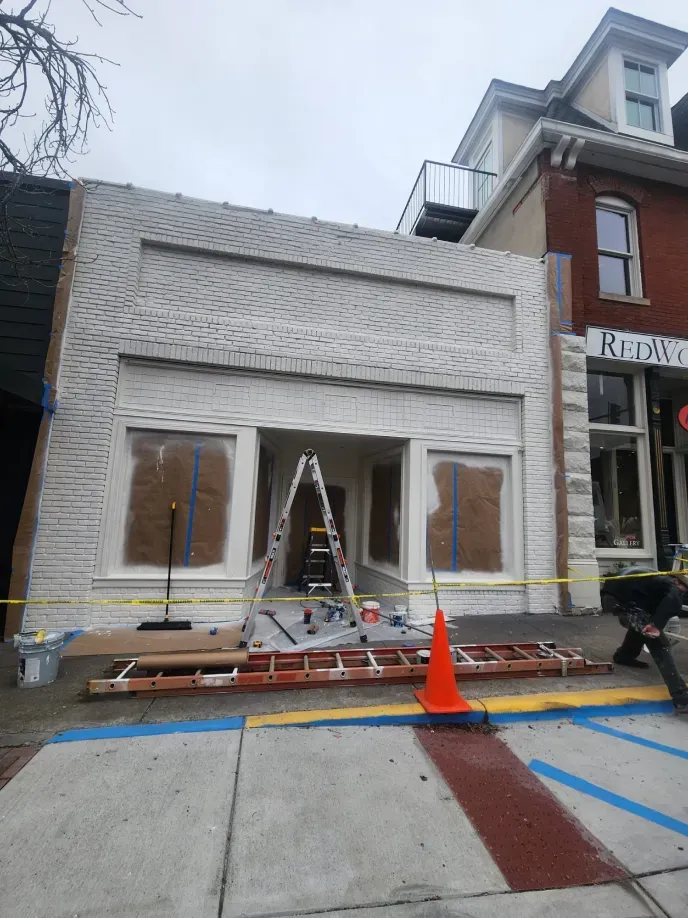 Building exterior being renovated, painted white. A-frame ladder inside doorway. Orange traffic cone in the foreground.