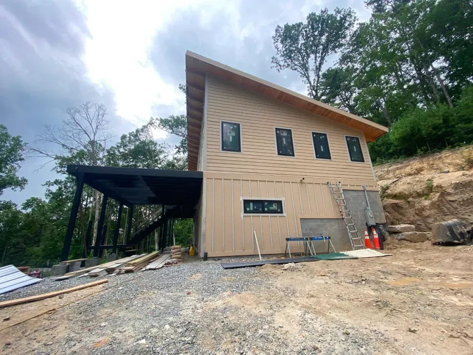Modern two-story house with angled roof and carport, under construction on a hillside, surrounded by trees.