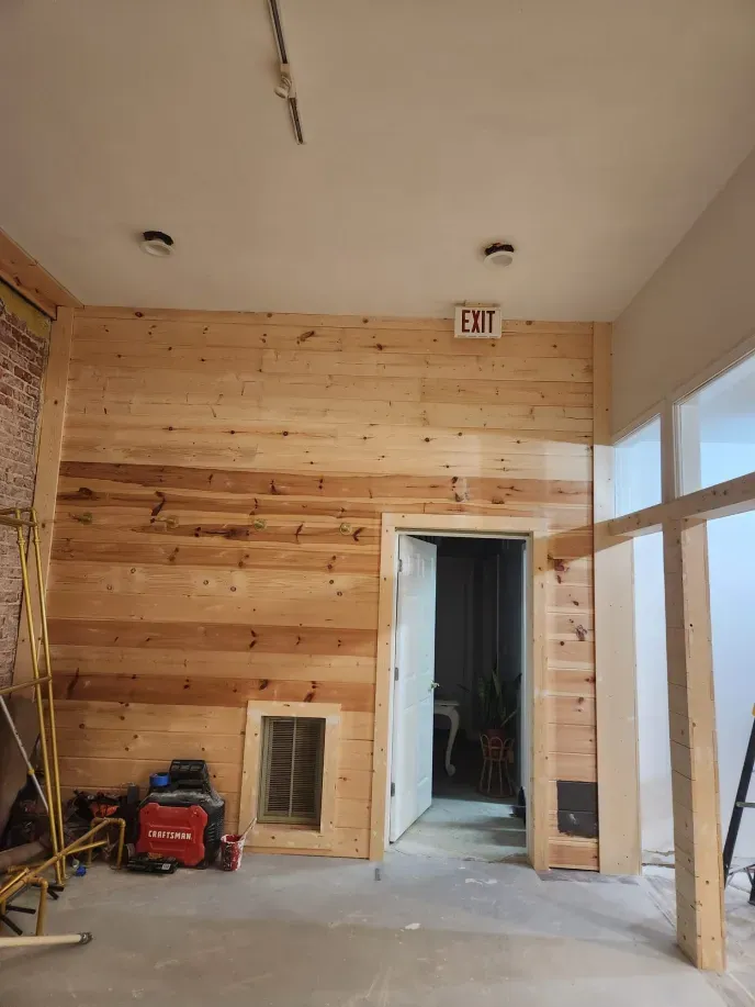 Interior with wood-paneled wall, exit sign, door, unfinished framing, and tools on the floor.