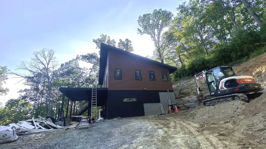 Modern cabin construction on a hillside. Brown siding, black accents, Bobcat excavator. Sunny day.