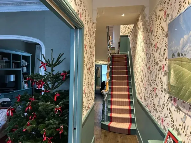 Hallway with floral wallpaper, a red-carpeted staircase, and a Christmas tree visible through a doorway.