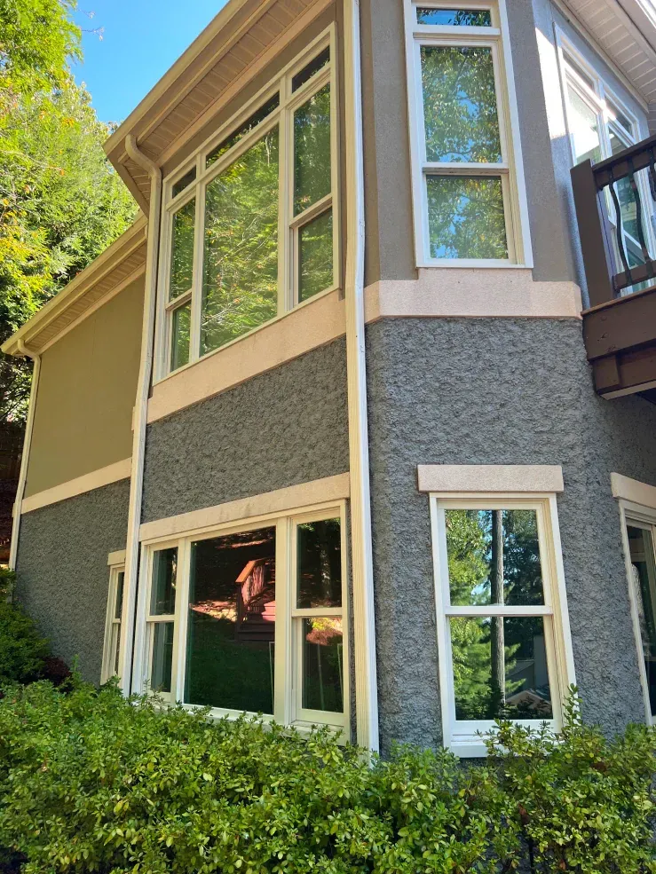 Two-story house exterior with large windows, stucco walls, and tan trim. Green foliage in foreground.