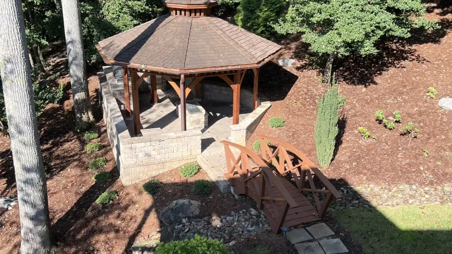 Wooden gazebo with a bridge over a dry creek bed, in a wooded area. Brown mulch.