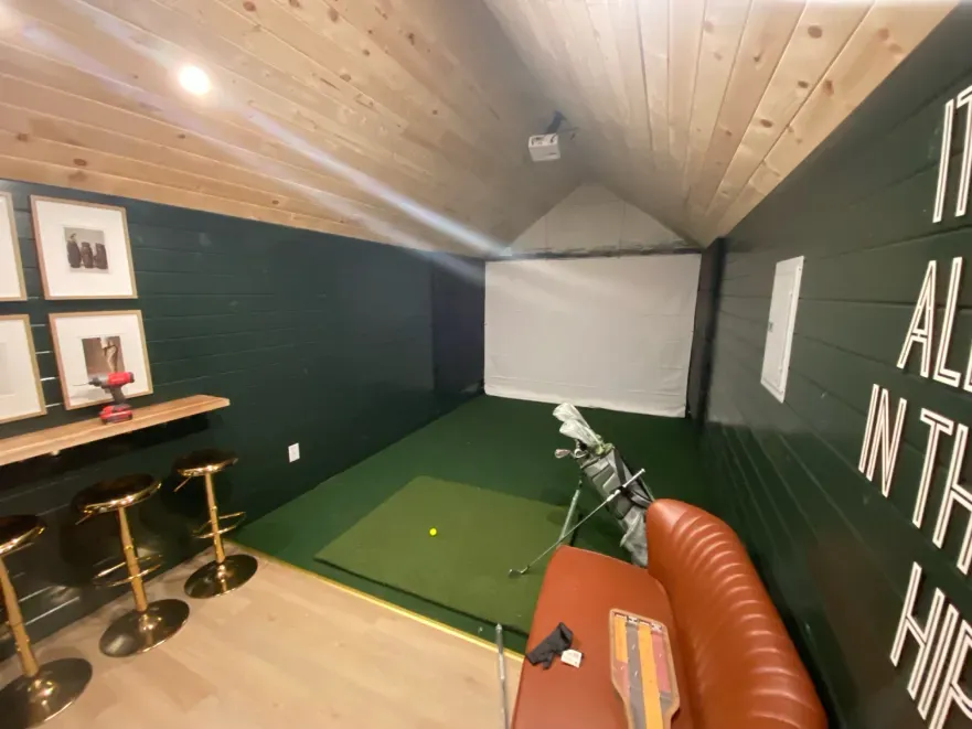 Golf simulator room with green walls, wooden ceiling, screen, golf mat, and bar seating.