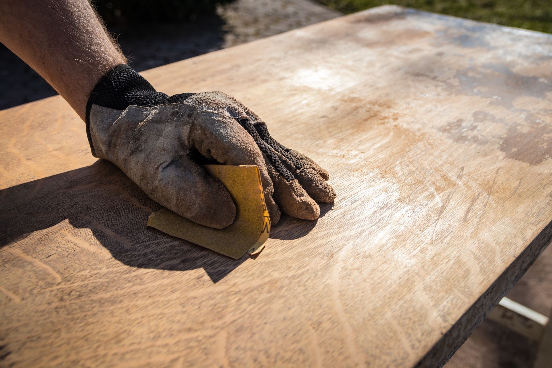 Gloved hand sanding a wooden surface with a piece of sandpaper.