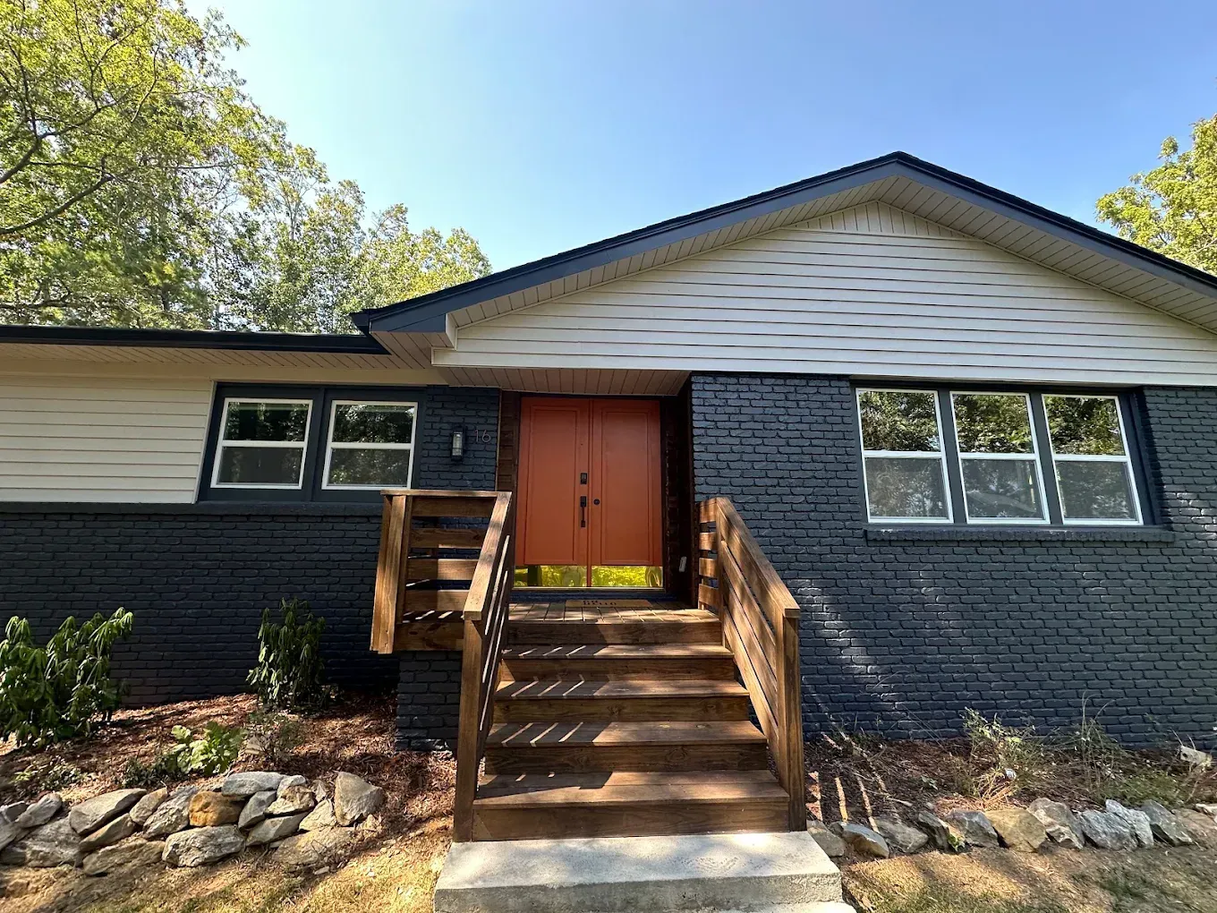 Dark gray brick house with orange front door, wooden steps and railing, windows, and trees.