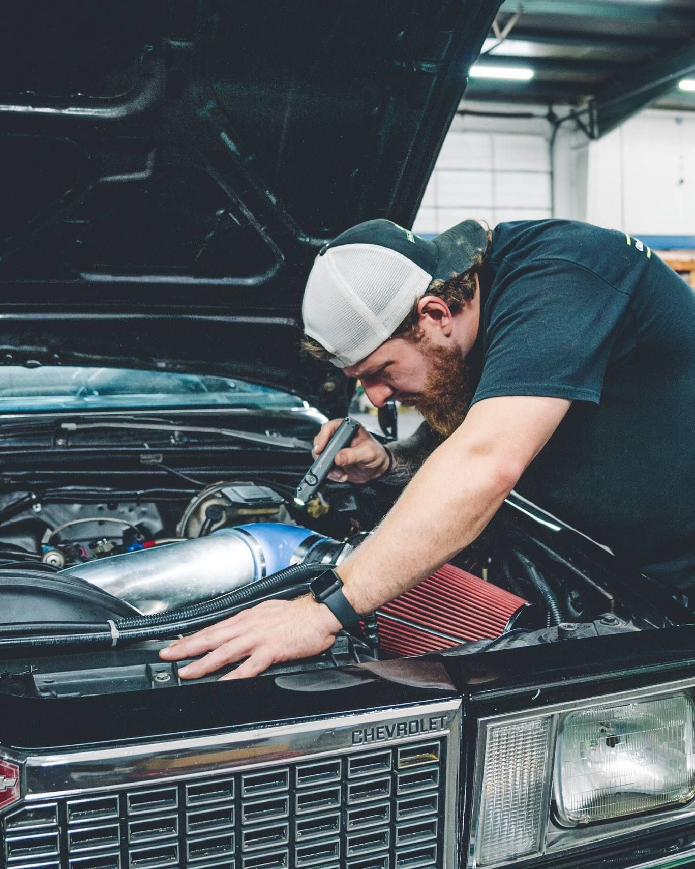 Mechanic examining a car engine. He's holding a light and leaning over the hood. | SK Mechanic