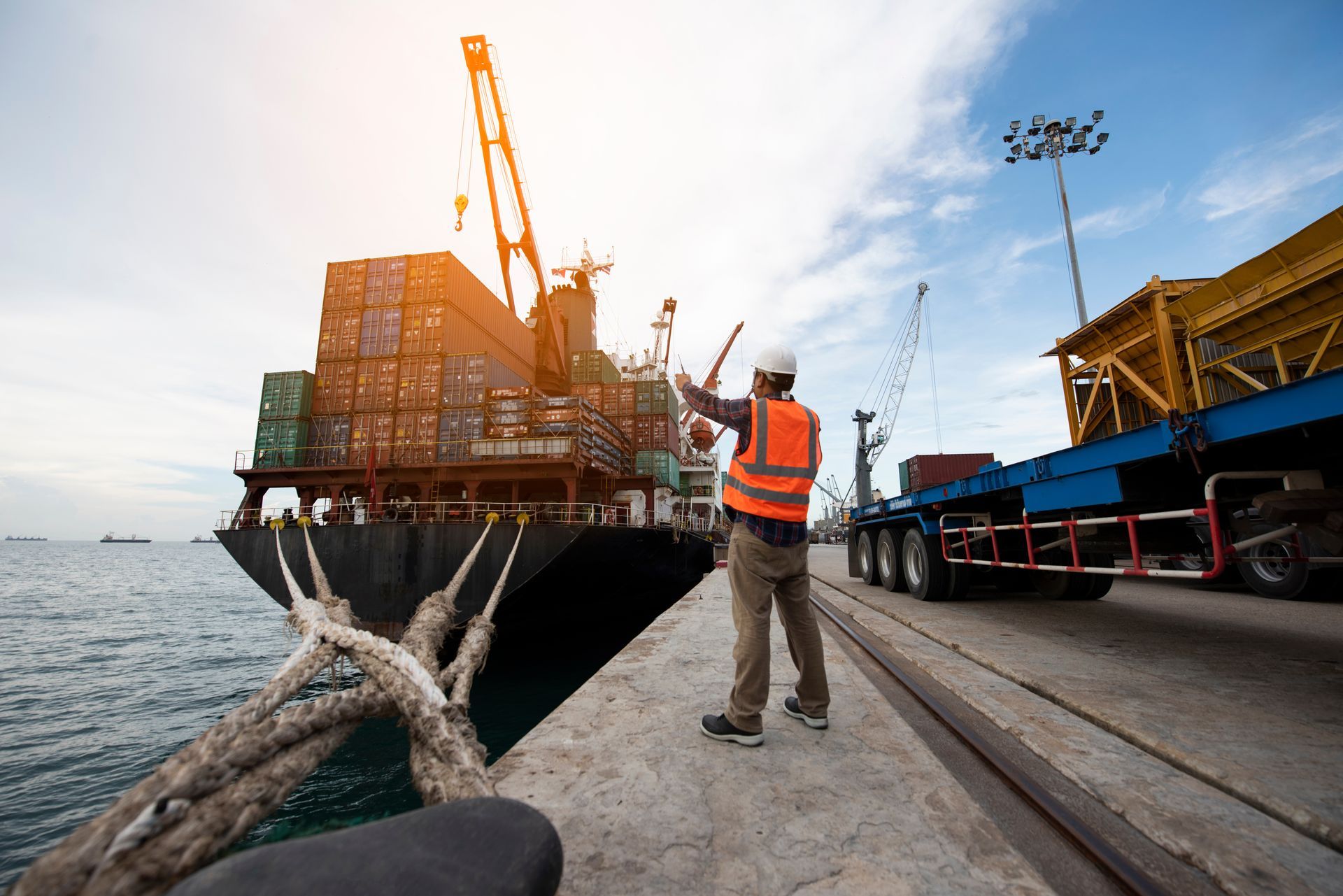 Cargo ship being loaded at a dock, with a worker in an orange vest signaling near the crane. Cargo ship being loaded at a dock, with a worker in an orange vest signaling near the crane.