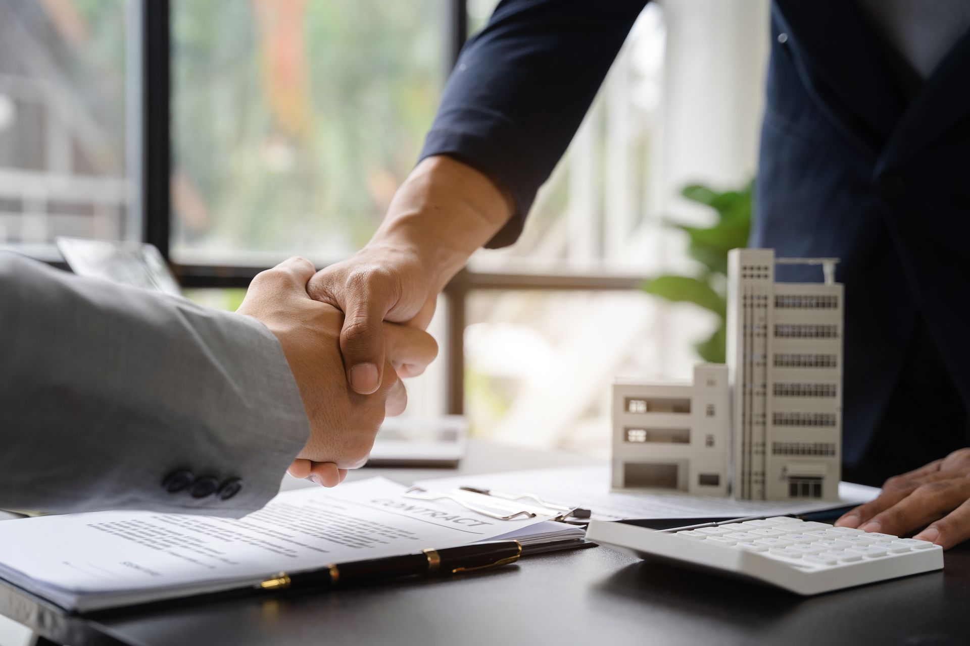 Businesspeople shaking hands over a desk with documents, calculator, and model building in an office Businesspeople shaking hands over a desk with documents, calculator, and model building in an office