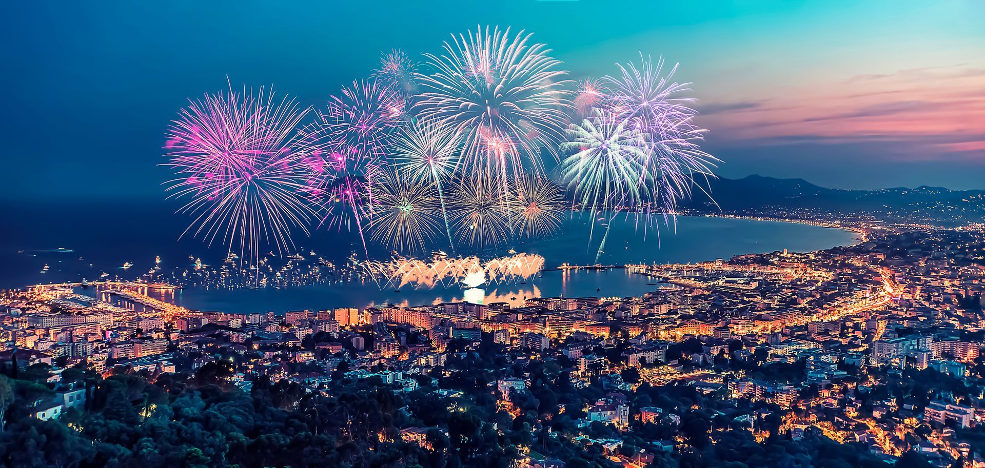 Fireworks exploding over a city at night with a bay. Pink, white, and purple fireworks.