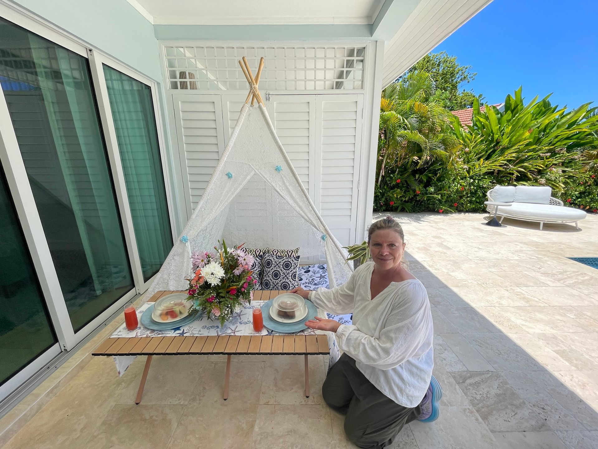 Woman kneeling by decorated table under a tent, with food and flowers. Tropical setting.