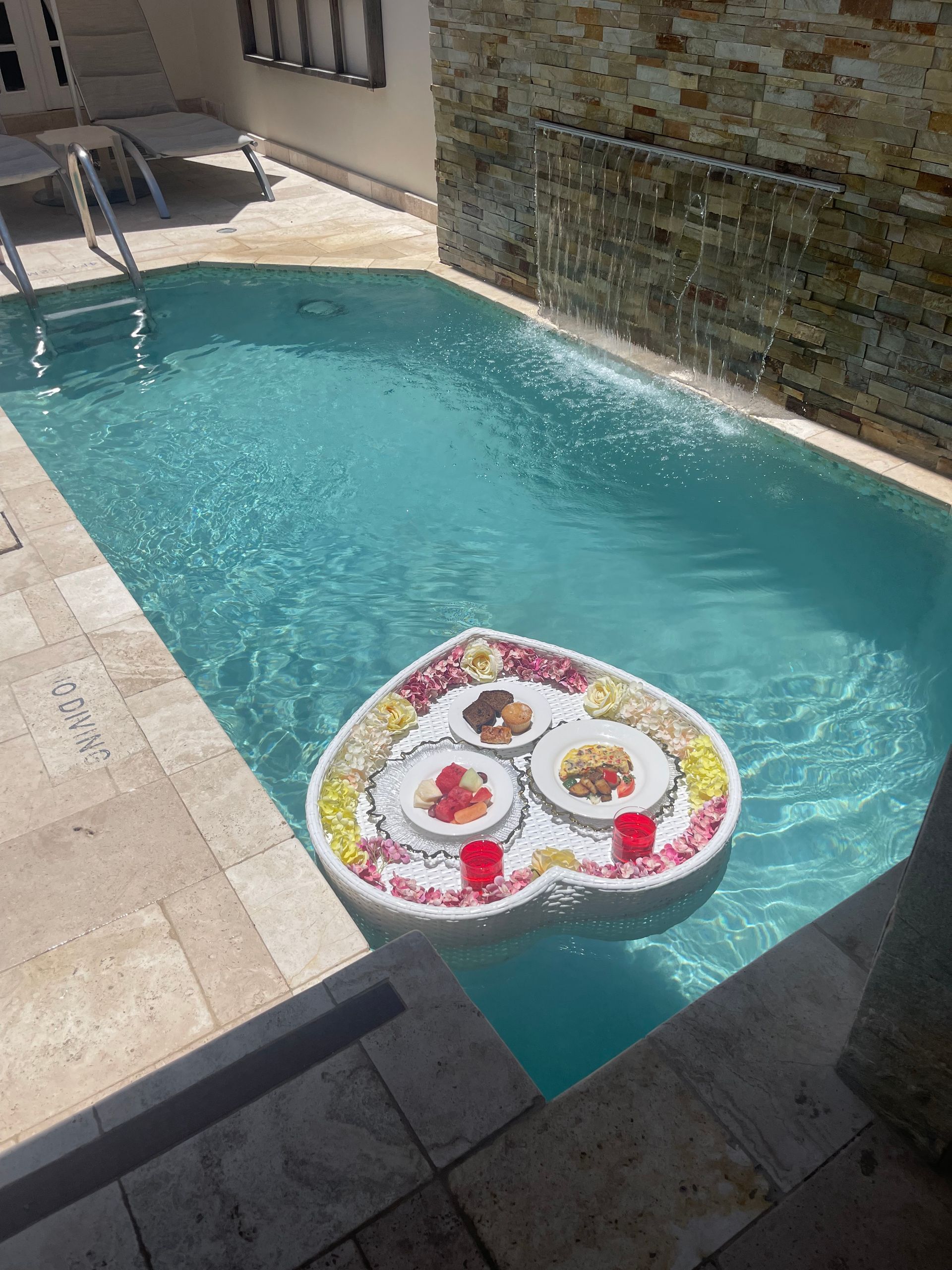 Heart-shaped floating tray with food and drinks in a pool, near a waterfall and beige stone.