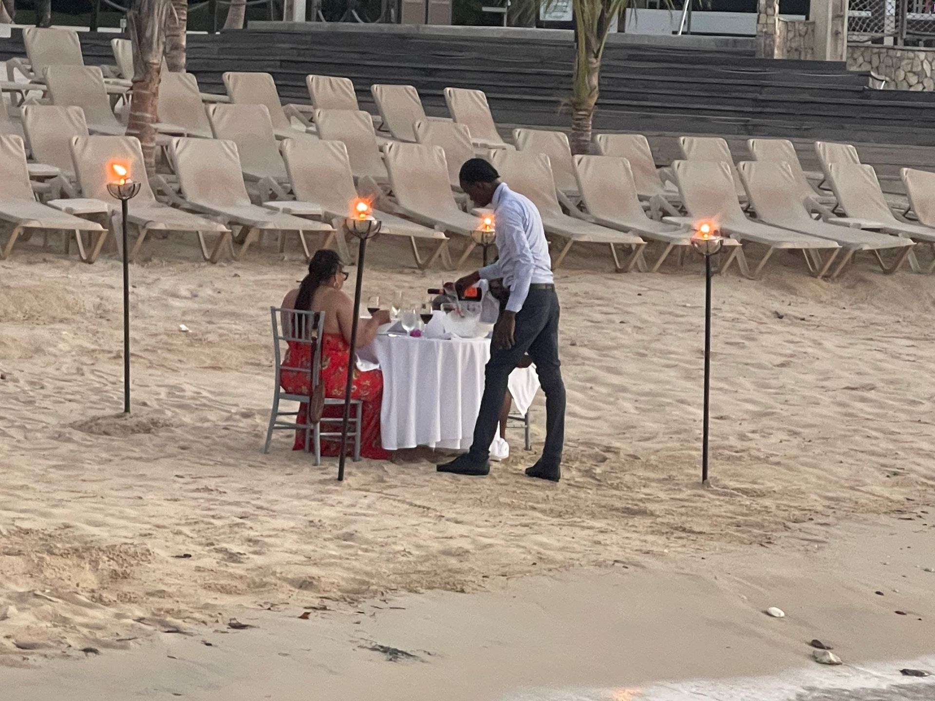 Man serving a woman at a beachside table set for a romantic dinner. Torch lights, beach chairs in the background.