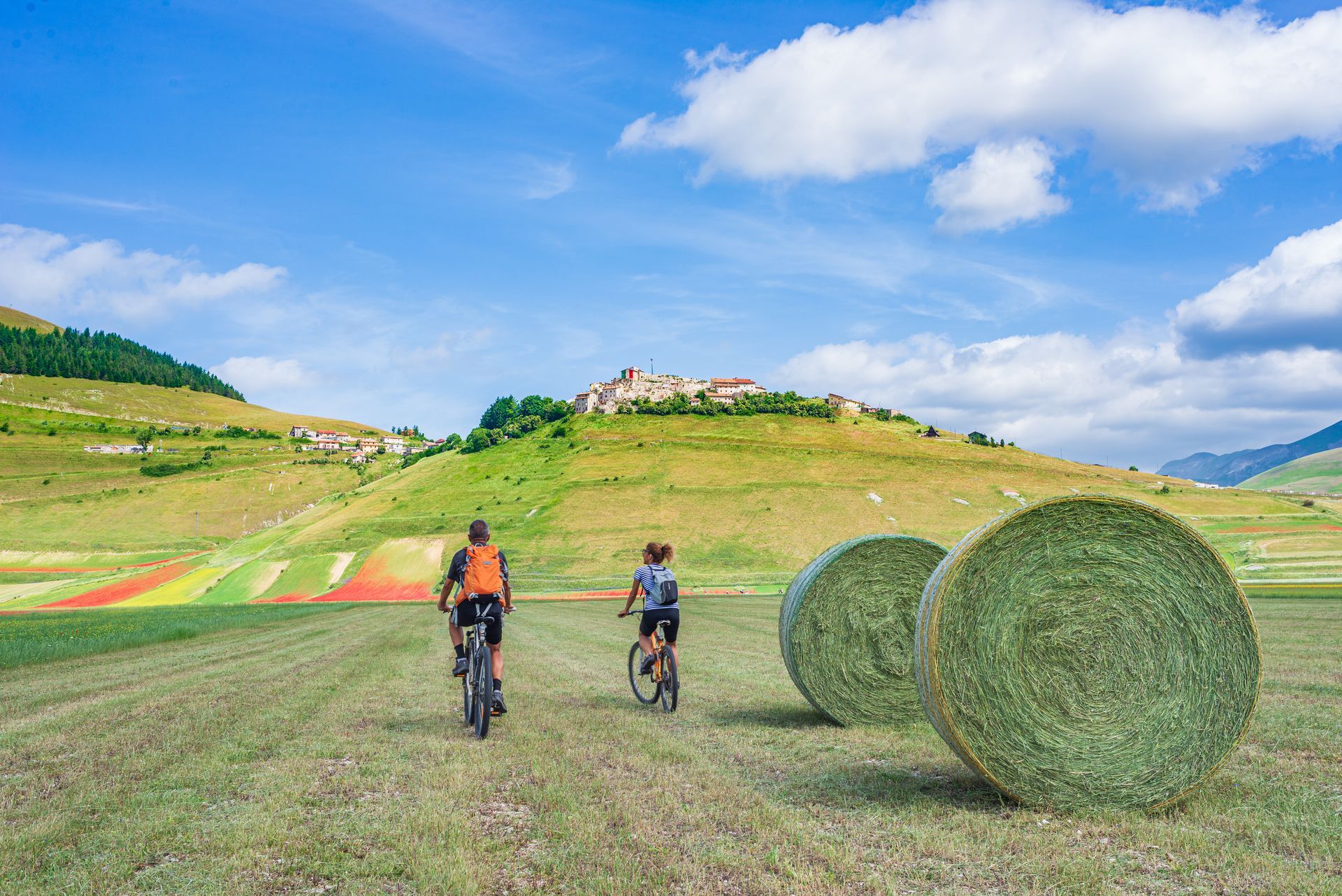 Two people are riding bicycles in a field next to bales of hay. COPYRIGHT USED WITH PERMISSION