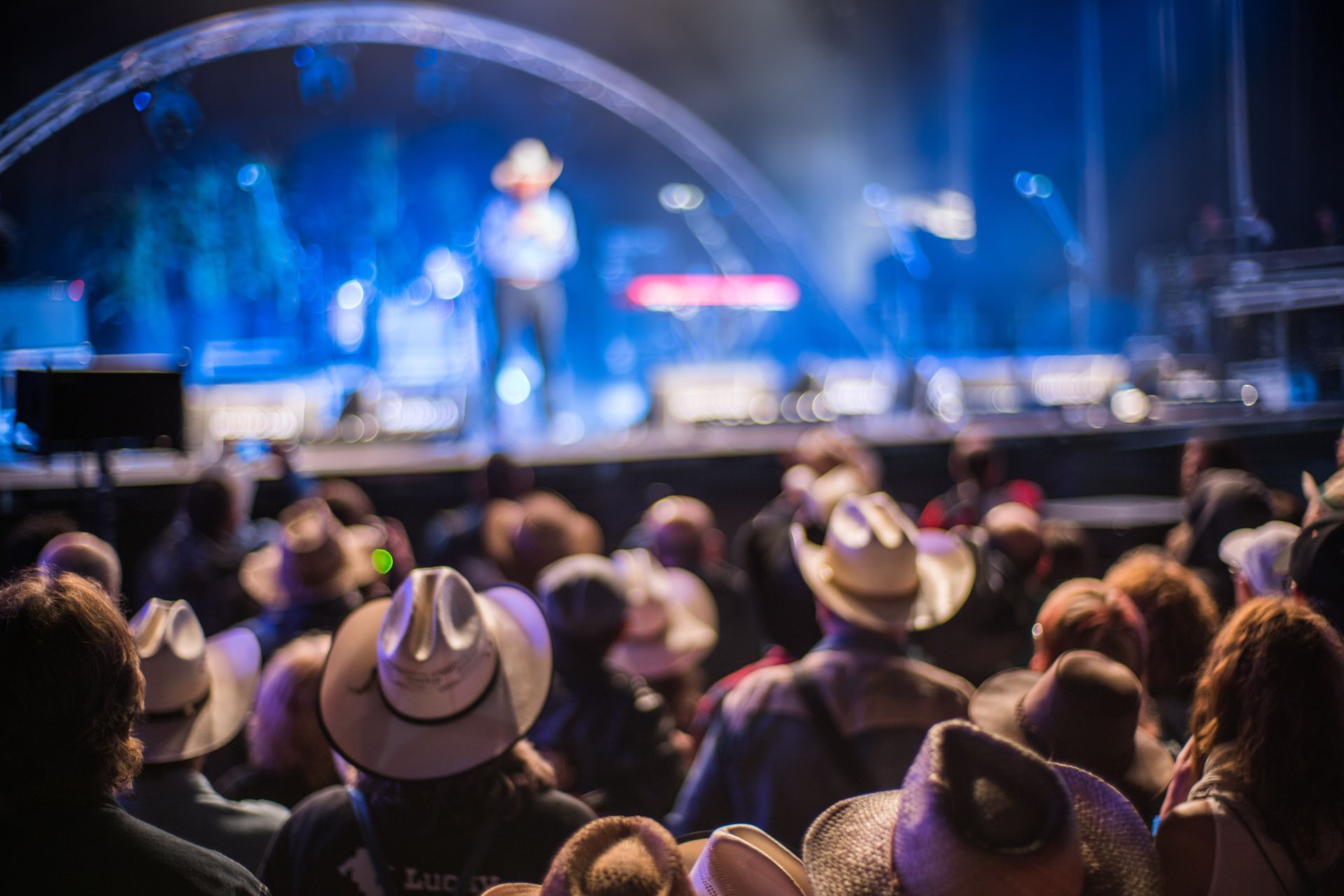 Audience at a country music concert, watching a performer on a brightly lit stage; many wear cowboy hats.