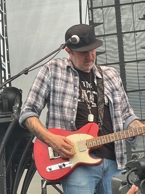Man playing a red electric guitar on stage, wearing a black hat, plaid shirt, and jeans.