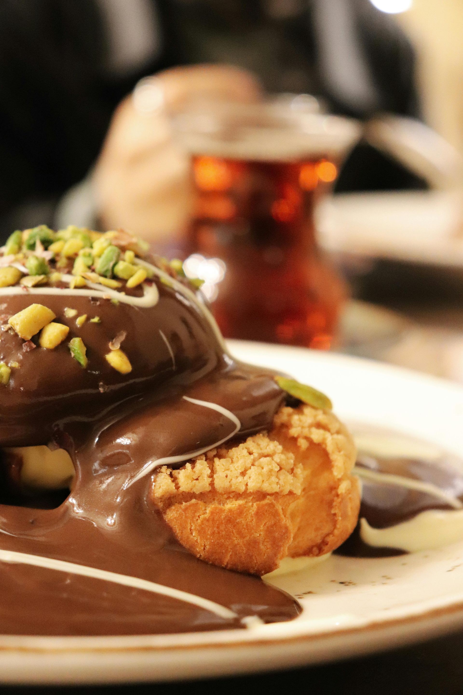 Profiteroles with chocolate sauce and pistachio garnish on a plate, tea in background.