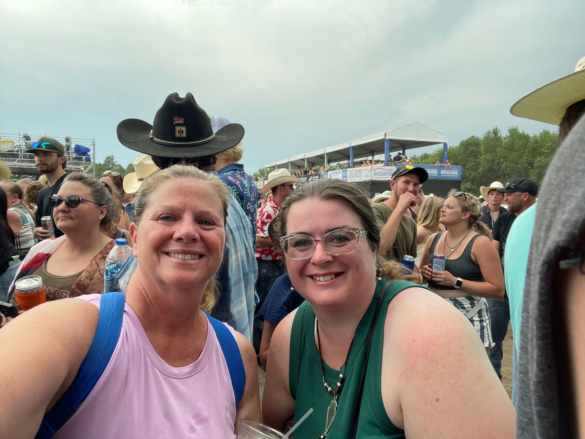 Two women smile at the camera at an outdoor event with many people, cowboys hats and drinks.