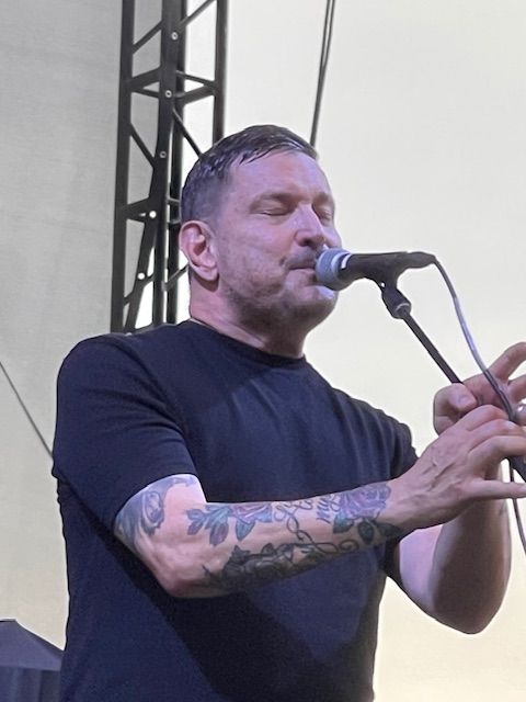 Man singing into a microphone on stage, arm tattoos visible, wearing a black shirt.