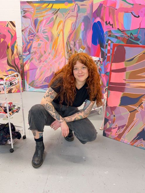 Woman with red hair in studio, squatting, smiling. Surrounded by colorful abstract paintings.