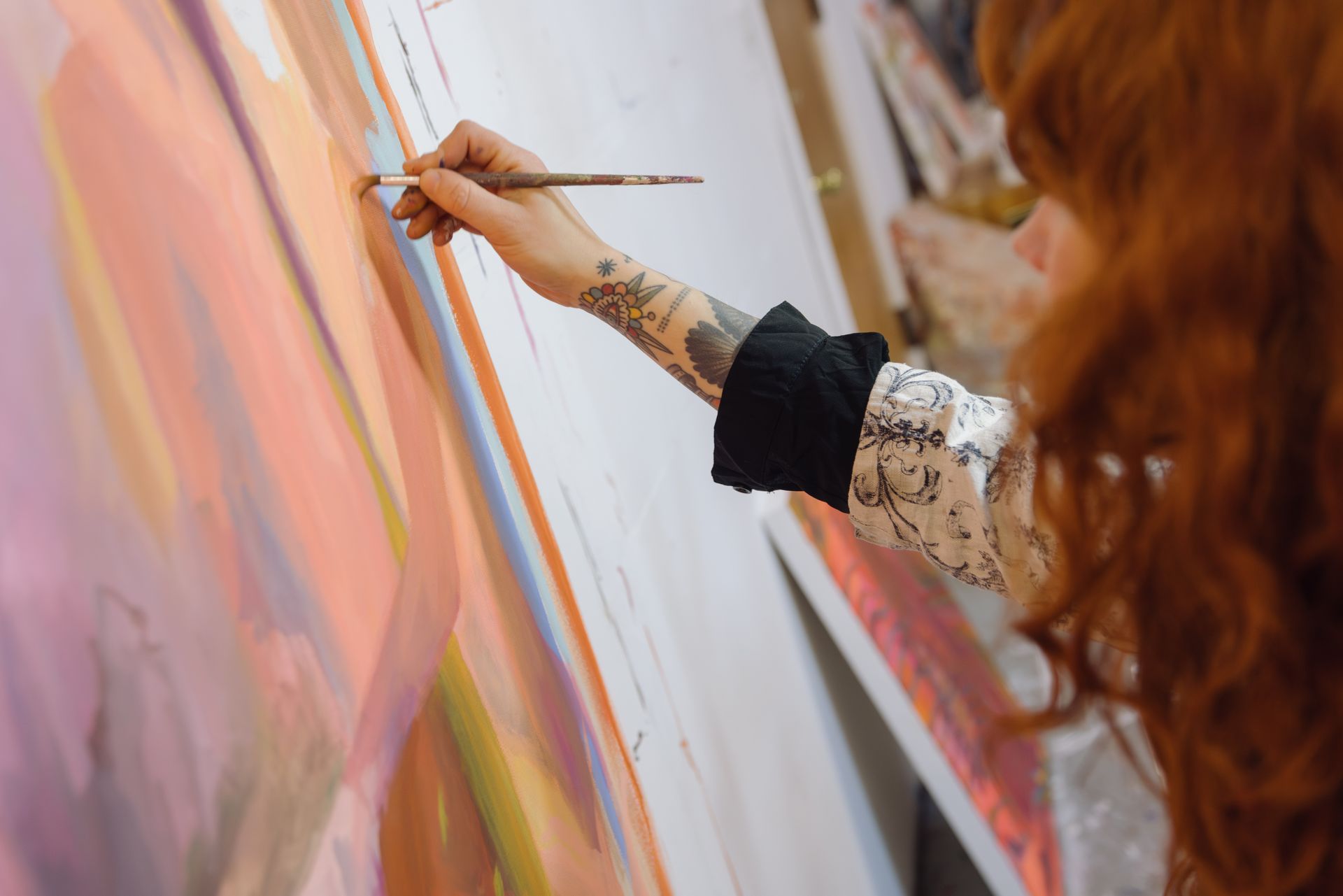Emily Wise, woman with red hair and tattoos painting on a large canvas, using a brush.