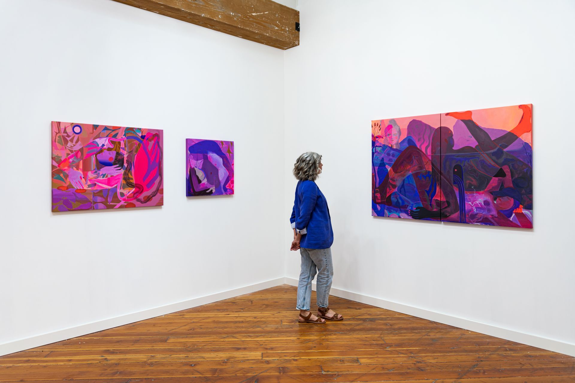 A person looks at vibrant abstract paintings in a white-walled gallery with a wooden floor.