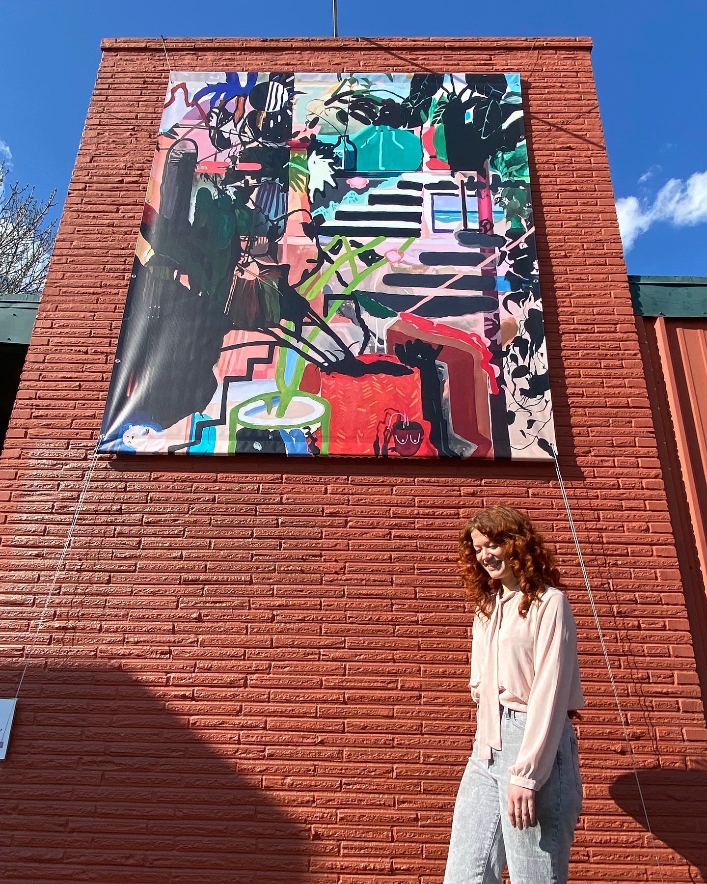 Emily Wise smiling next to a large, colorful abstract painting hanging on a red brick wall.
