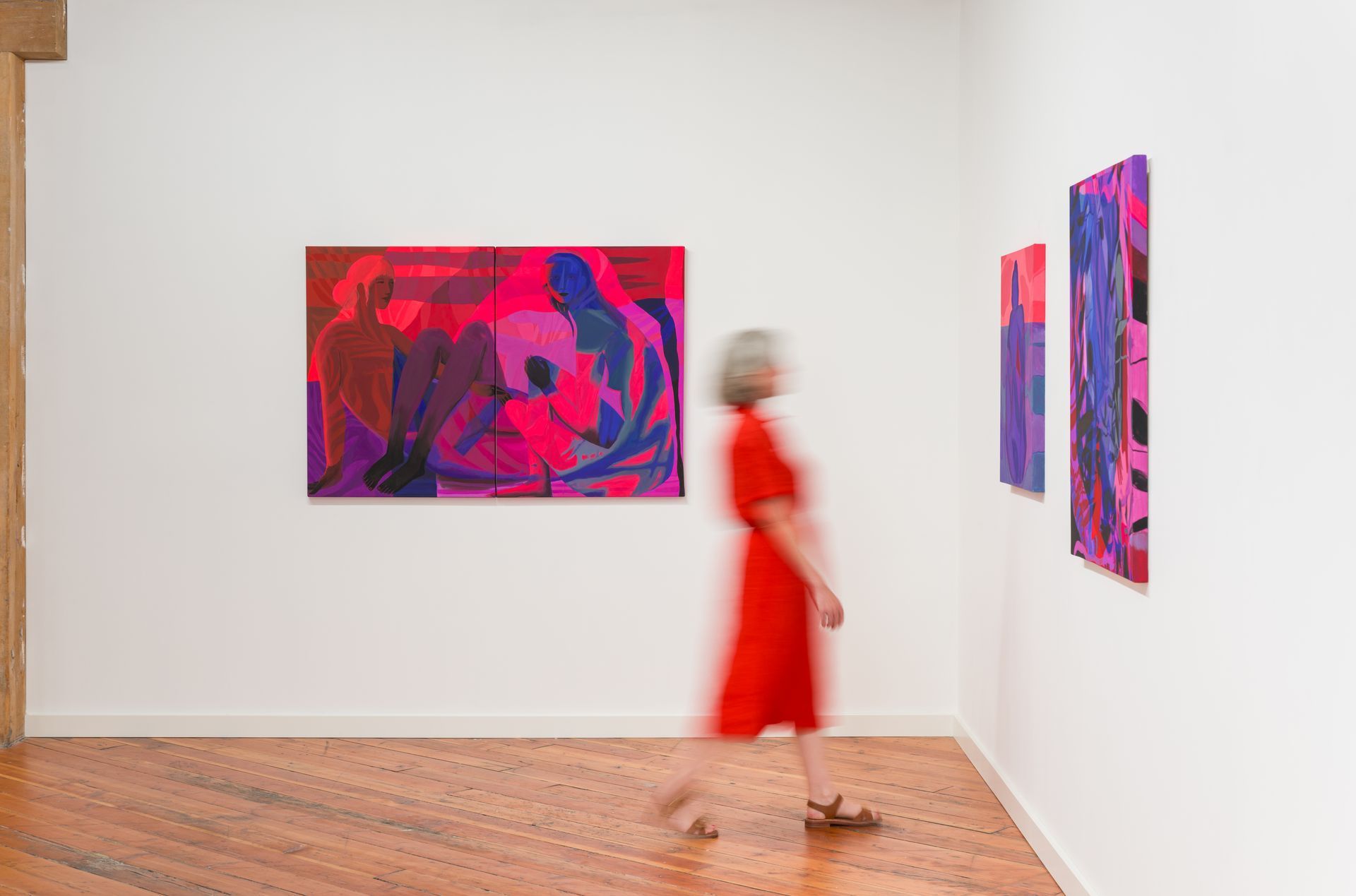Woman in red dress walks past paintings in a gallery; vibrant pinks, purples on white walls.