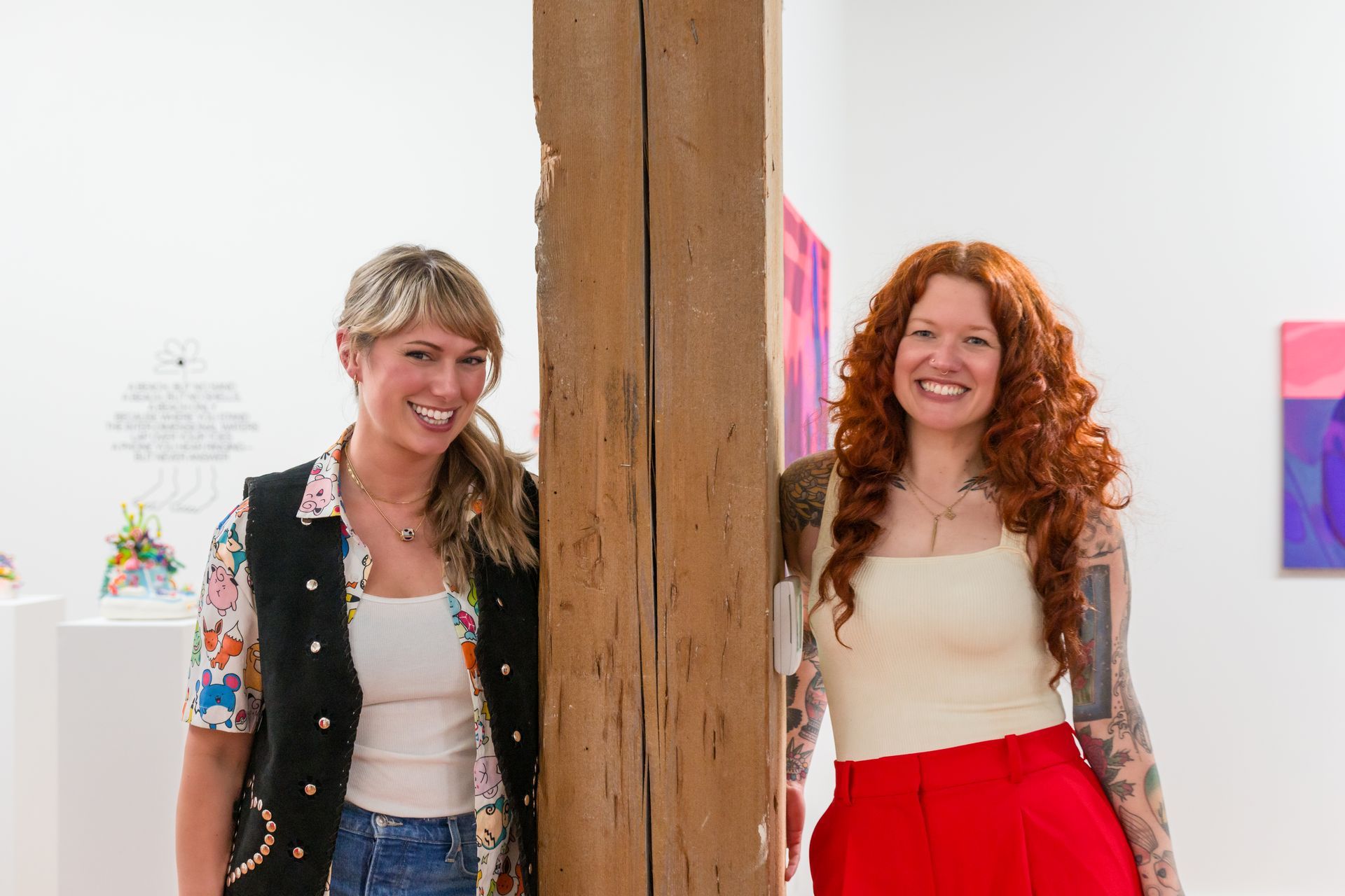 Emily Wise and another artist smile at the camera, posing by a wooden pillar in a gallery with colorful art.