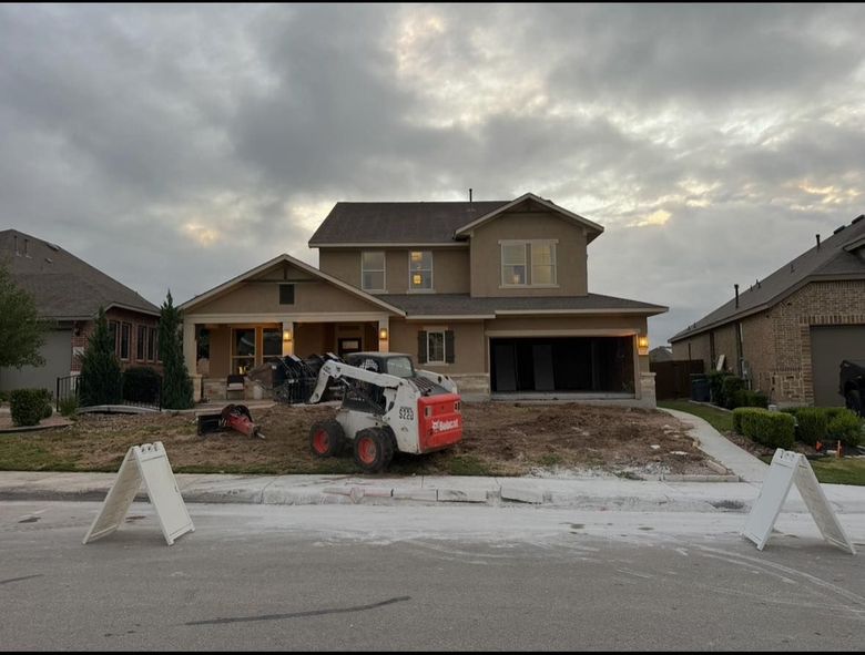 A two-story house with a bobcat in the yard during a landscaping project, under a cloudy sky.