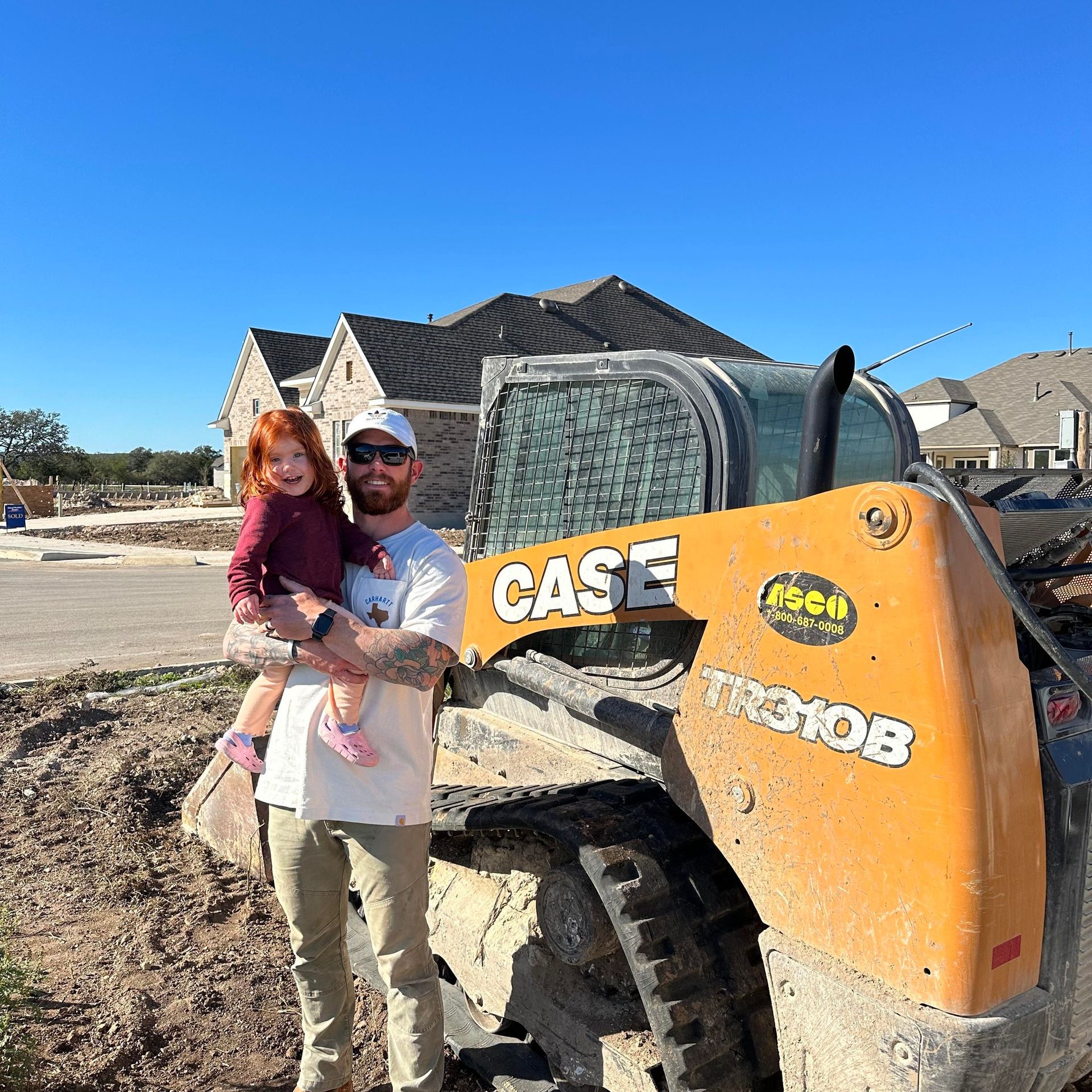 Man holding a red-haired child poses in front of an orange Case skid steer on a construction site; houses in background.