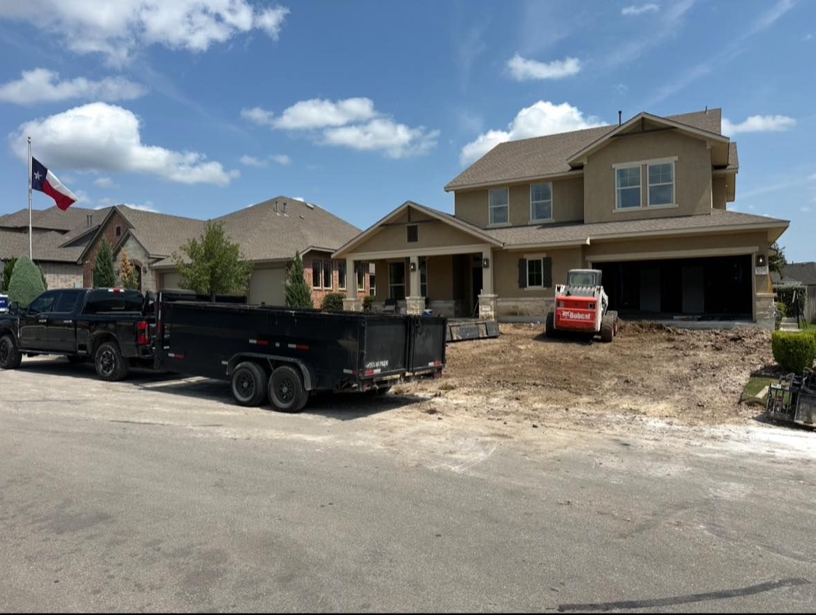 House under construction, black truck, trailer, bobcat, and Texas flag on a sunny day.