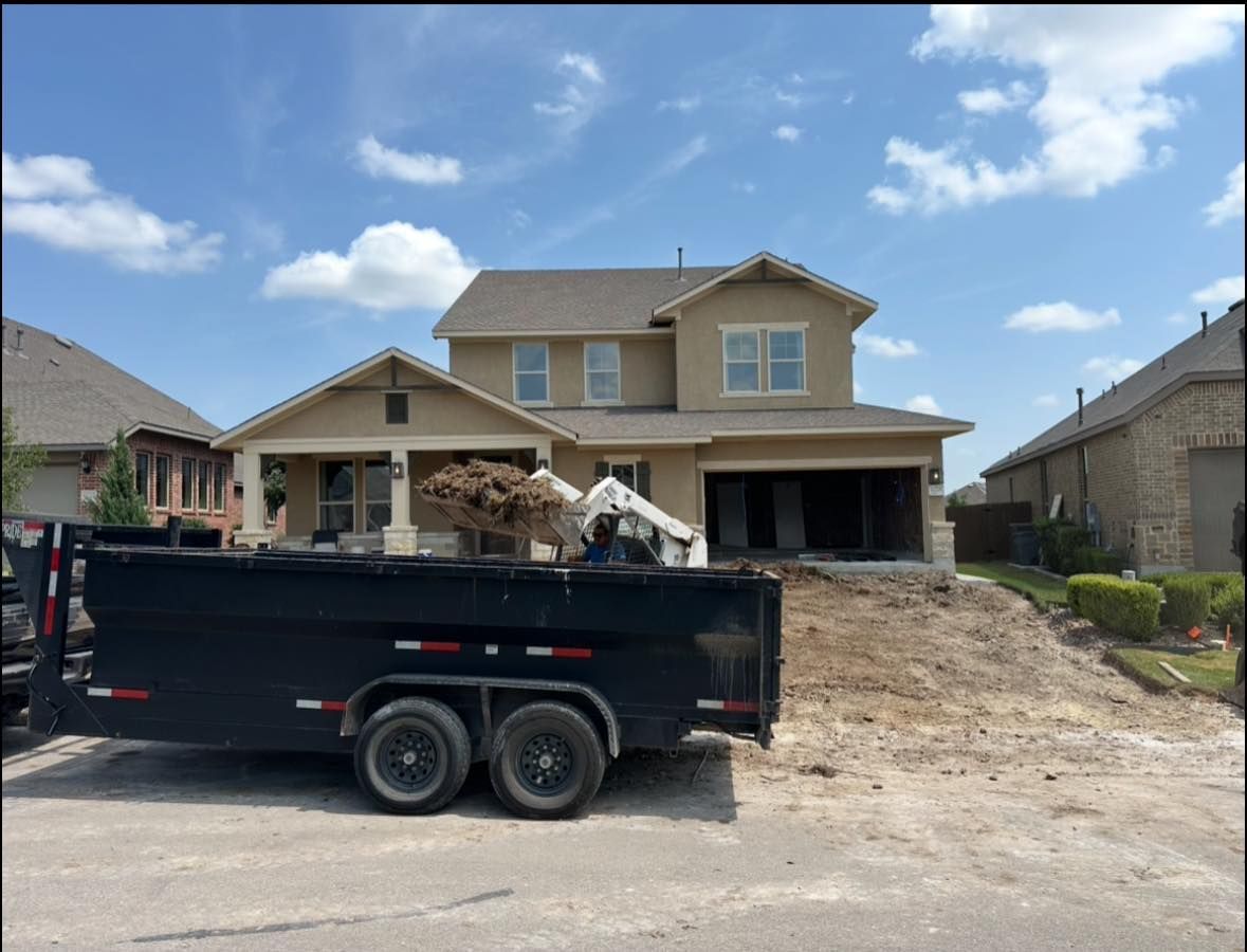 A two-story beige house with a dirt mound and trailer, possibly under construction, on a sunny day.