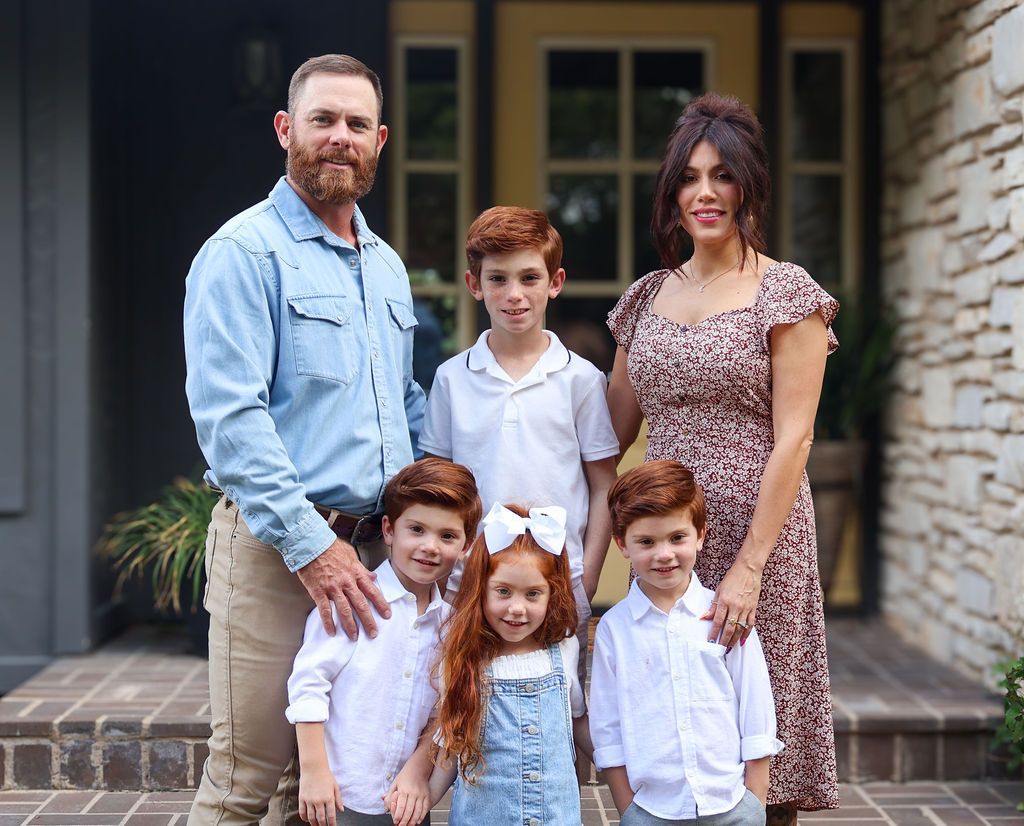 Man holding a red-haired child poses in front of an orange Case skid steer on a construction site; houses in background.