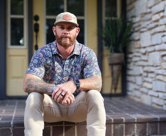 Man with beard and tattoos sits on steps in front of a house, wearing a cap and floral shirt.