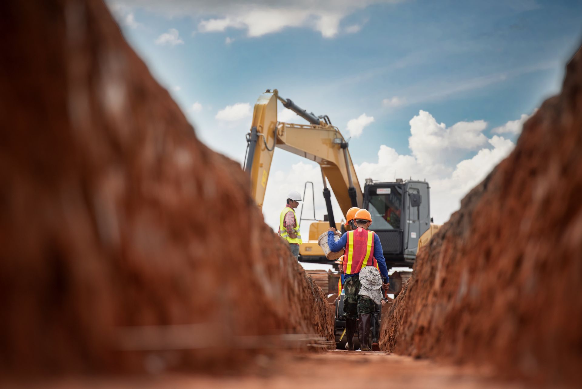 Construction workers inspect a deep trench, excavator in the background, under a blue sky.