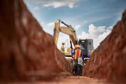 Construction workers inspect a deep trench, excavator in the background, under a blue sky.