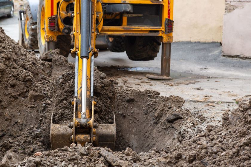 Yellow backhoe digging a trench in the dirt on a street near a building.